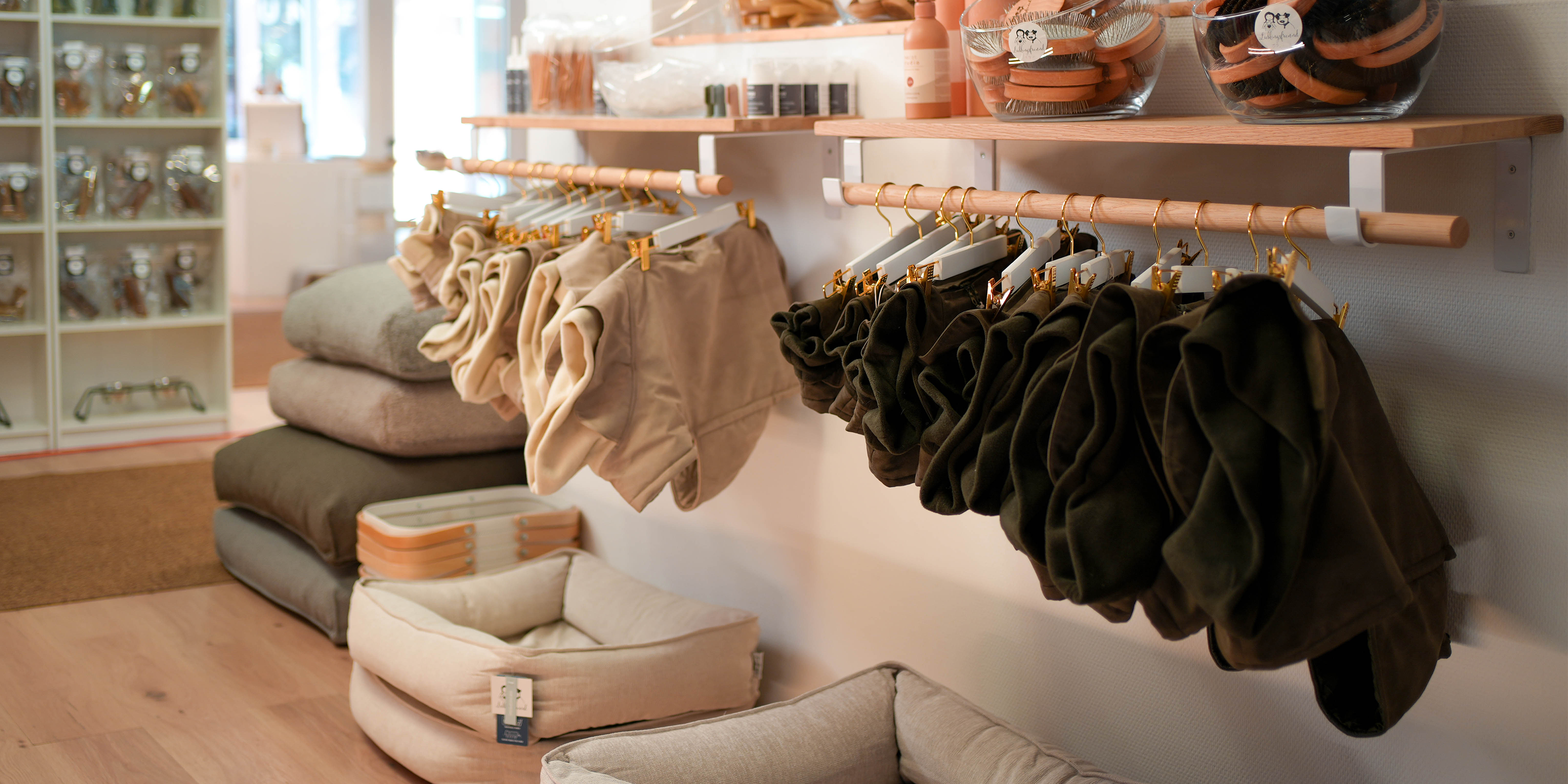 Nachhaltige Hundebekleidung in Beige und Olivgrün, sorgfältig auf Kleiderbügeln präsentiert, daneben gemütliche Hundebetten und Accessoires in der Lieblingsfreund Boutique.